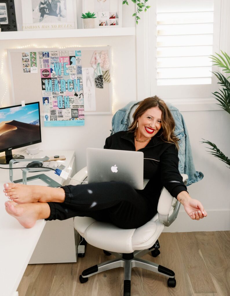 Ashley Freeborn. Profile photo of Ashley Freeborn, sitting with her feet up on a desk, smiling and holding her Macbook computer.