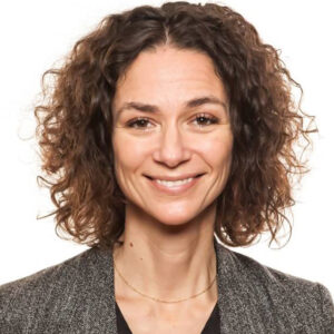 Julie Angus. Headshot of Julie Angus wearing a grey jacket standing in front of a bright white background.
