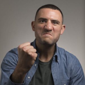 Paul Bissonnette. Headshot of Paul Bissonnette wearing a blue collared shirt over a black T-shirt. He is posing in front of a grey gradient background and holding up his fist.