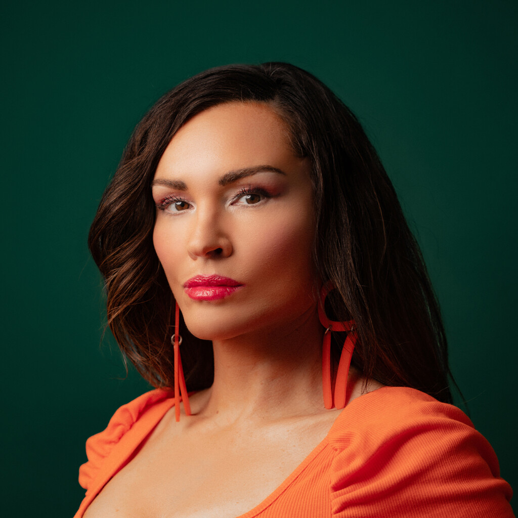 Angela Sterritt. Headshot of Angela Sterritt wearing an orange blouse and orange earrings. She is posing in front of a dark green back drop.