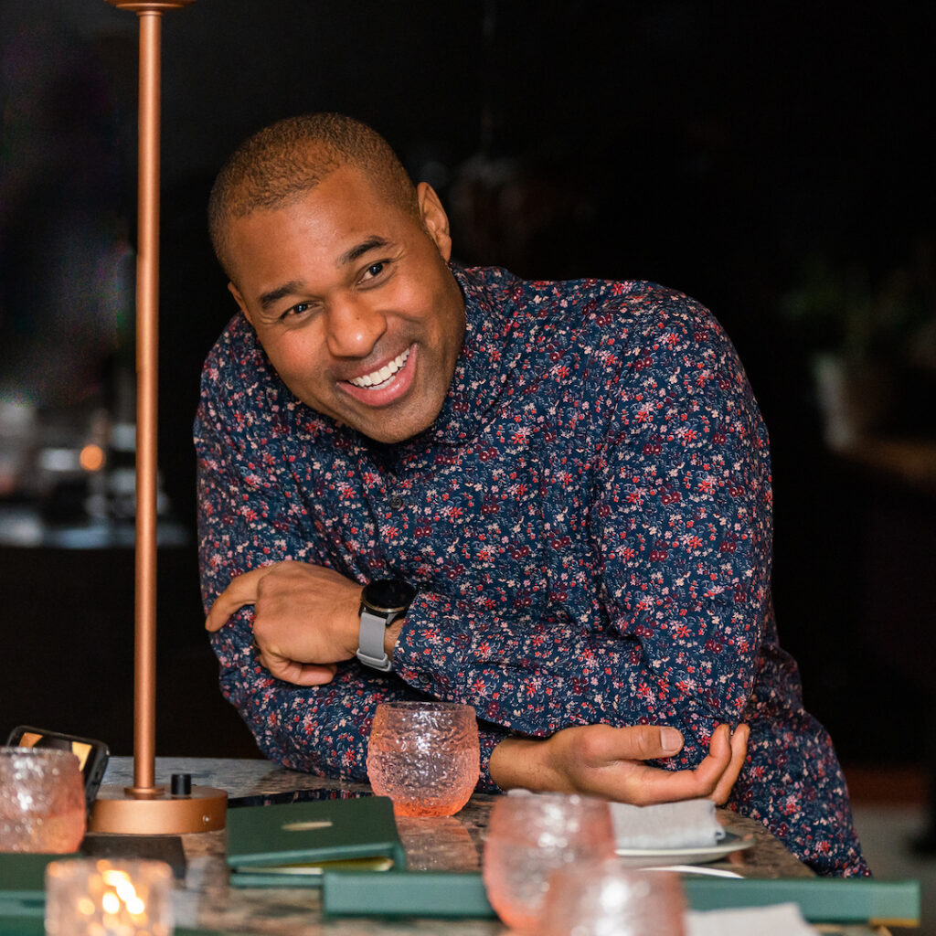 Cabral "Cabbie" Richards wearing a floral patterned shirt. He is smiling and leaning on his elbows on a table.