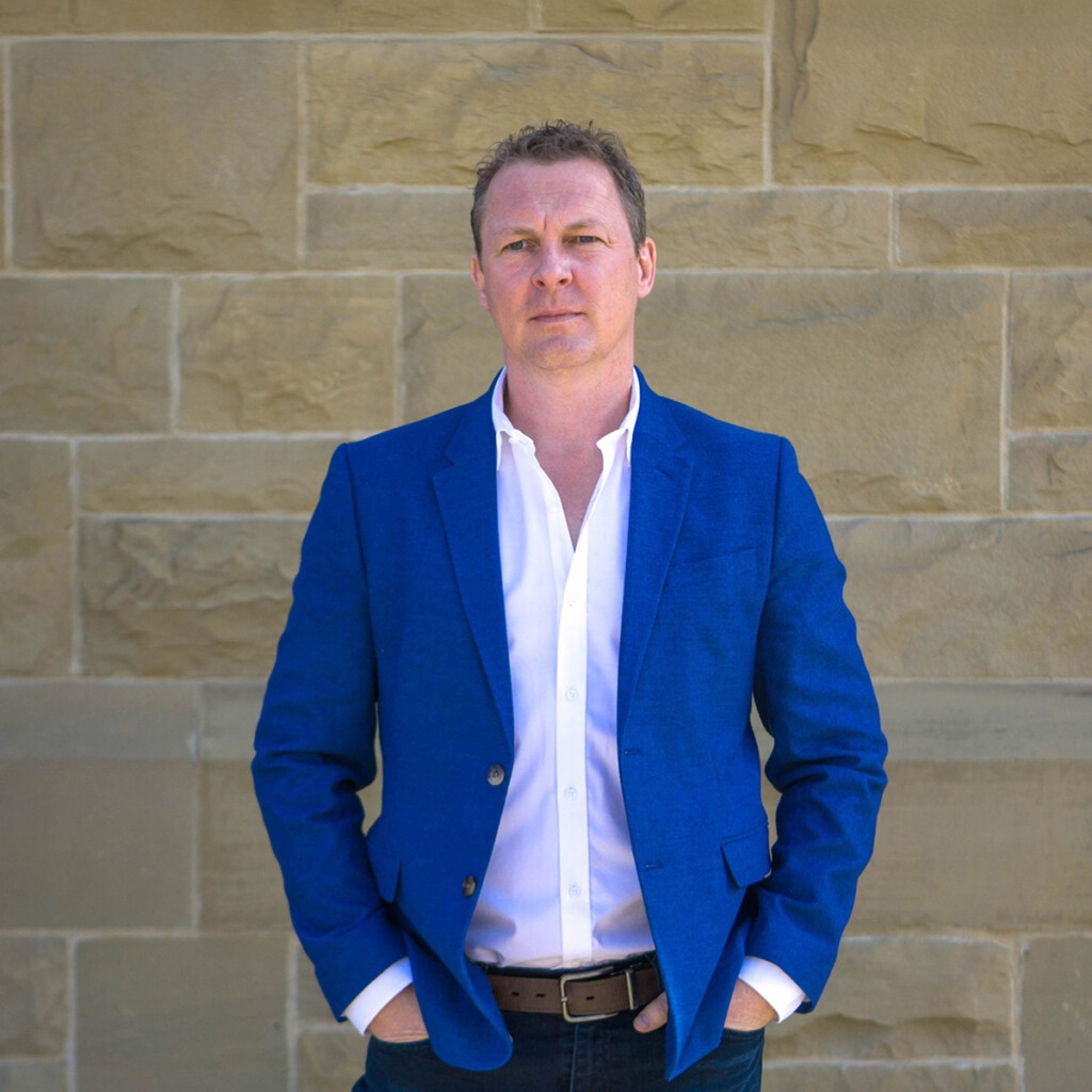 Doug Griffiths. Headshot of Doug Griffiths wearing a bright blue jacket over a white collared shirt. He is standing with his hands in his pockets in front of a brick wall.