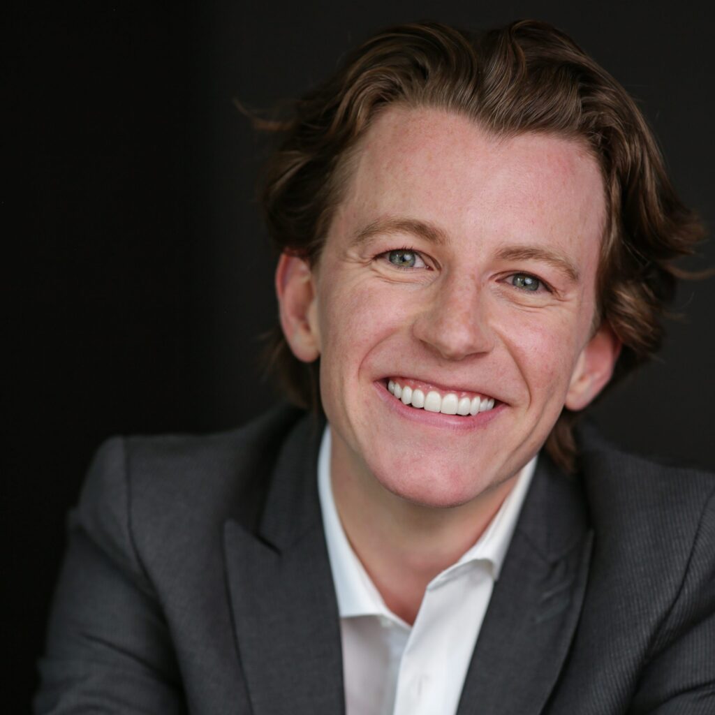 Harrison Browne professional headshot in front of a black background. He is smiling, wearing a white button up shirt underneath a dark grey blazer.
