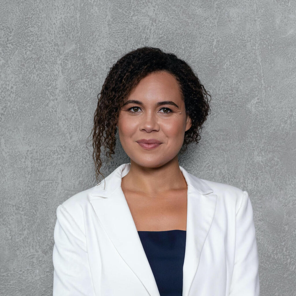 Iliana Oris Valiente headshot. Iliana is standing and smiling in front of a grey backdrop, wearing a black shirt underneath a white blazer.