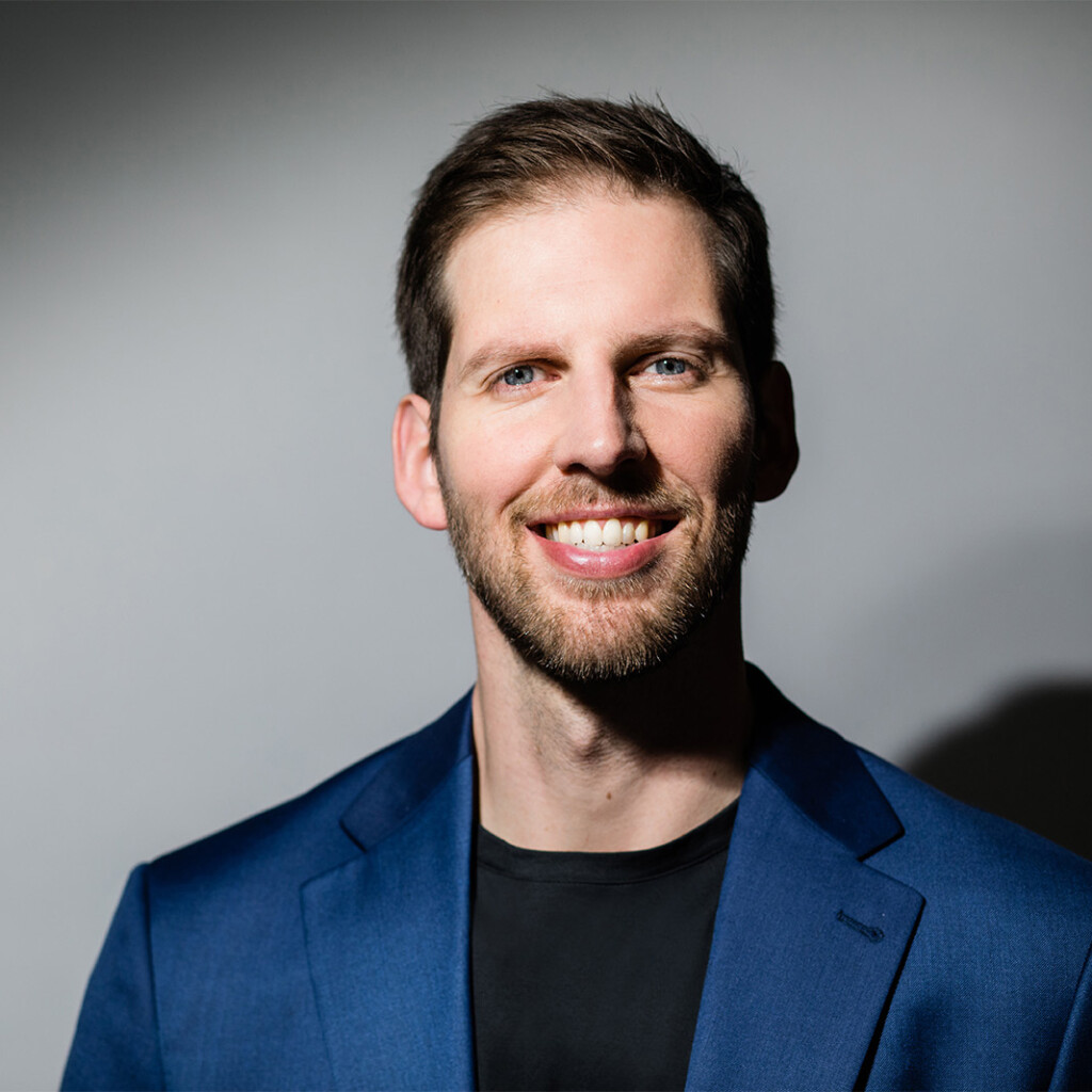 Jeremiah Brown. Headshot of Jeremiah Brown wearing a blue suit jacket over a black t-shirt. He is smiling and posing in front of a grey background.