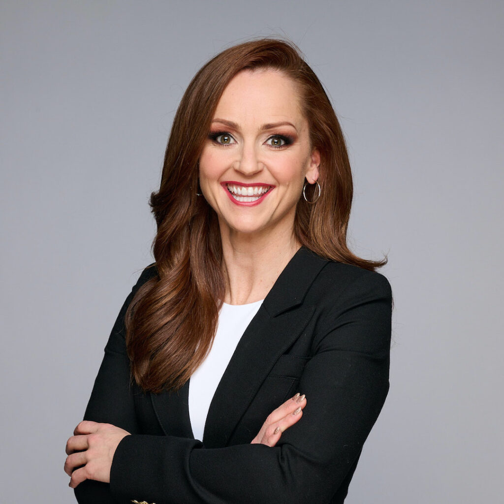 Headshot of Kate Beirness wearing a black jacket over a white top. He is smiling with her arms crossed and she's posing in front of a grey backdrop.