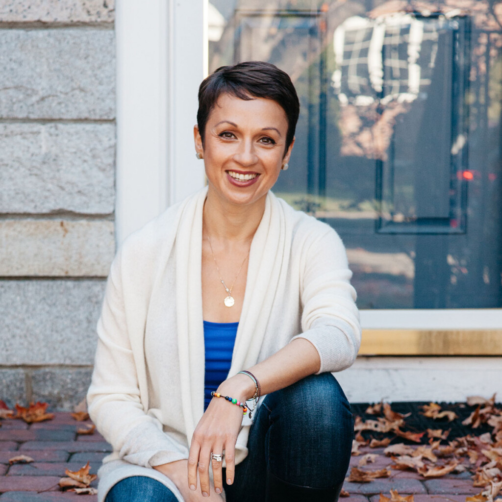Nataly Kogan. Photo of Nataly Kogan, sitting outside on the front steps out a house. She is wearing a white cardigan over a blue top with jeans.