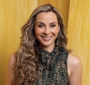 Andi Petrillo. Headshot of Andi Petrillo wearing a black and gold sleeveless top. She is smiling and posing in front of a yellow wall.