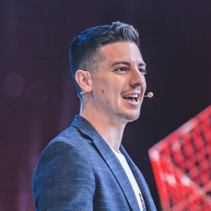 Shane Feldman. Headshot of Shane Feldman wearing a blue suit jacket over a white top. He's speaking into a headset microphone and standing sideways.