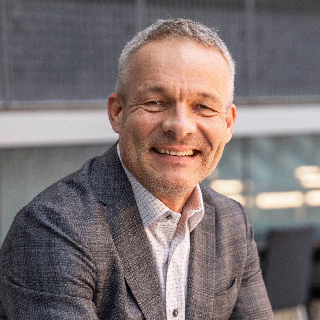 Urs Koenig. Headshot of Urs Hoenig, smiling, wearing a grew suit jacket over a white collared shirt posing in front of a window.