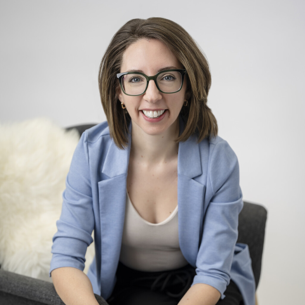 Ashley Margeson. Headshot of Ashley Margeson, wearing a light blue jacket over a white top, sitting sideways on a grey armchair. She is wearing glasses and smiling.