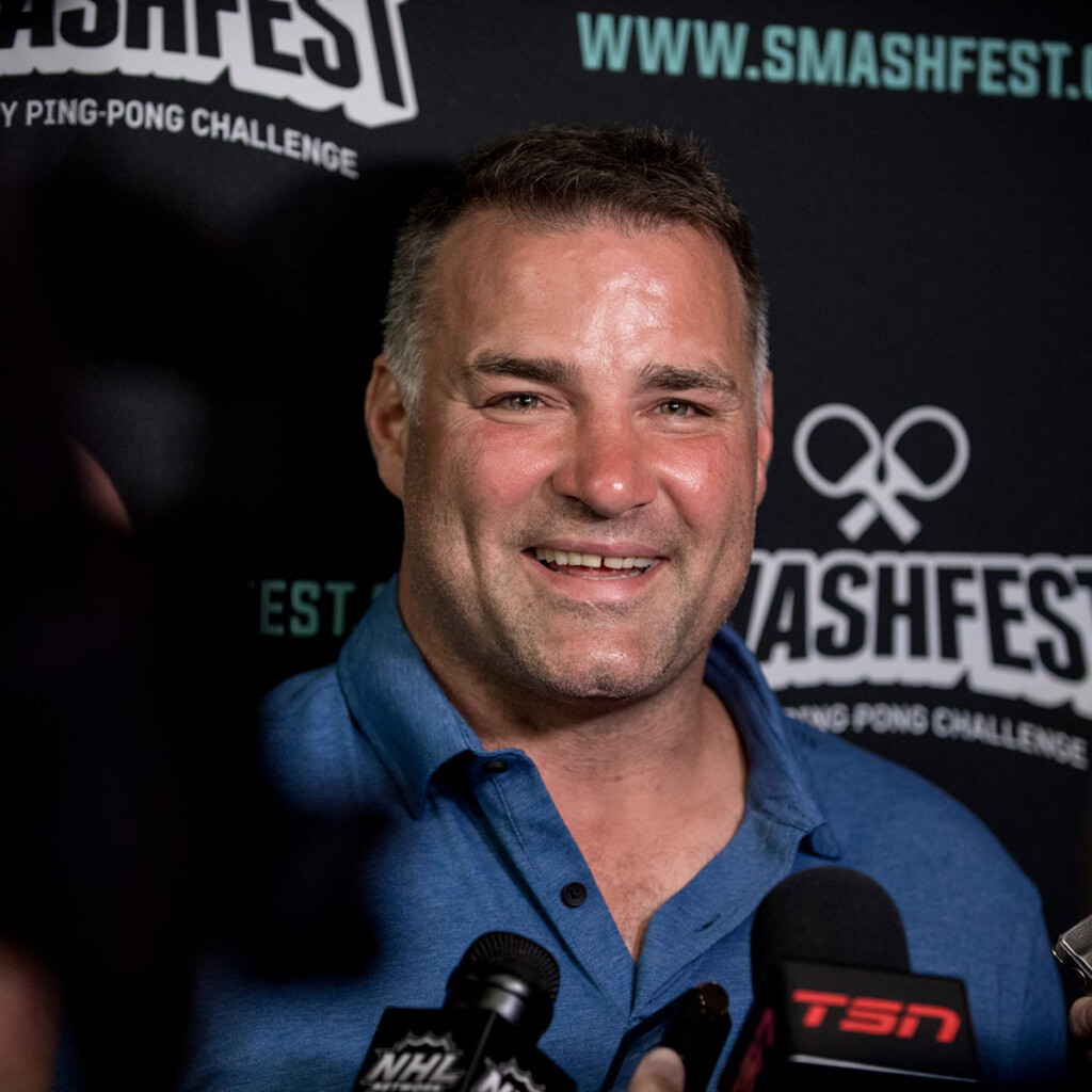 Eric Lindros. Headshot of Eric Lindros, smiling, wearing a blue collared shirt and standing in front of microphones.