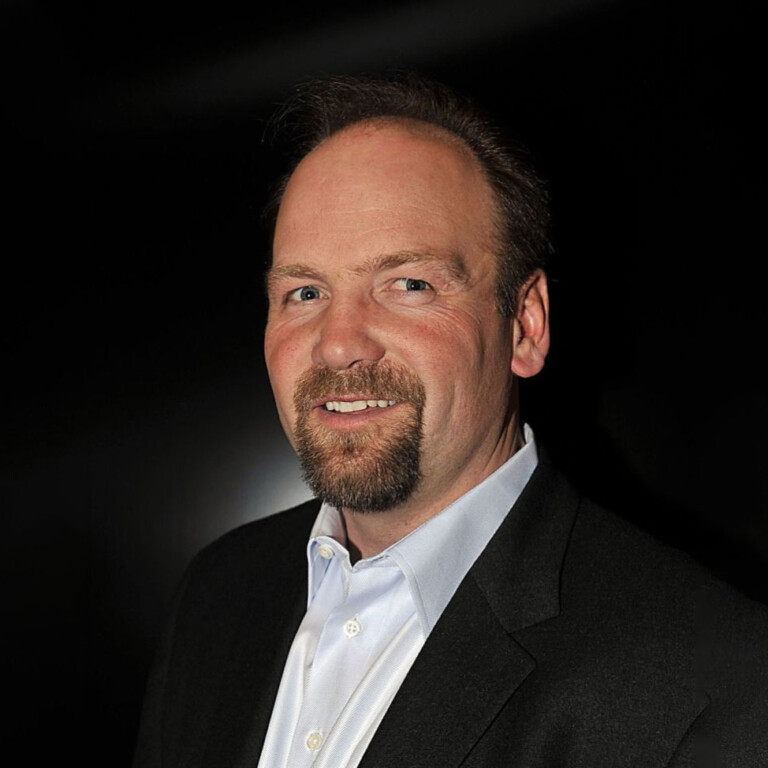 Wendel Clark. Headshot of Wendel Clark wearing a black suit jacket over a white collared shirt, posing in front of a black background.