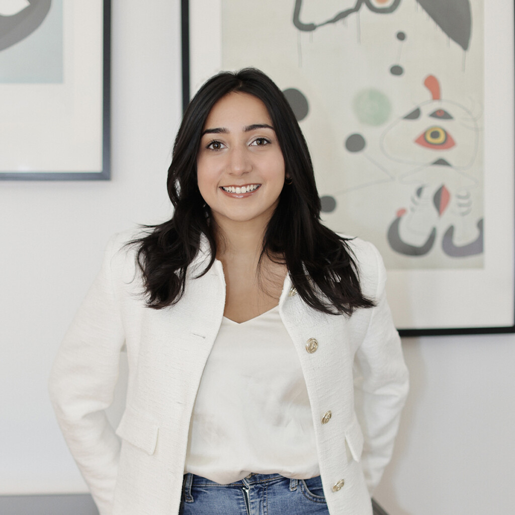 Inana Lalani. headshot of Inana Lalani wearing a white jacket over a white top. She is standing in front of a white wall with a framed piece of abstract art. She is smiling and has her hands in her pockets.