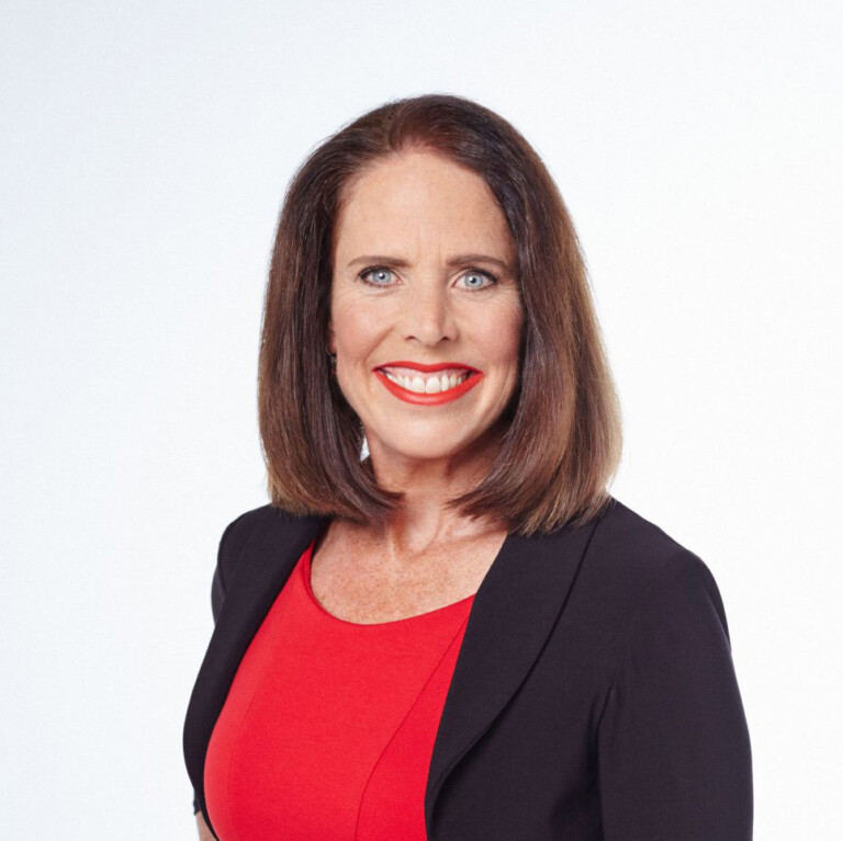 Meg Soper. Headshot of Meg Soper wearing a black jacket over a red top. She is smiling and standing in front of a plain white background.