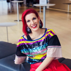 Professor Maja. Headshot or professor Maja wearing a colourful top and a red skirt. She is sitting down on a black bench.