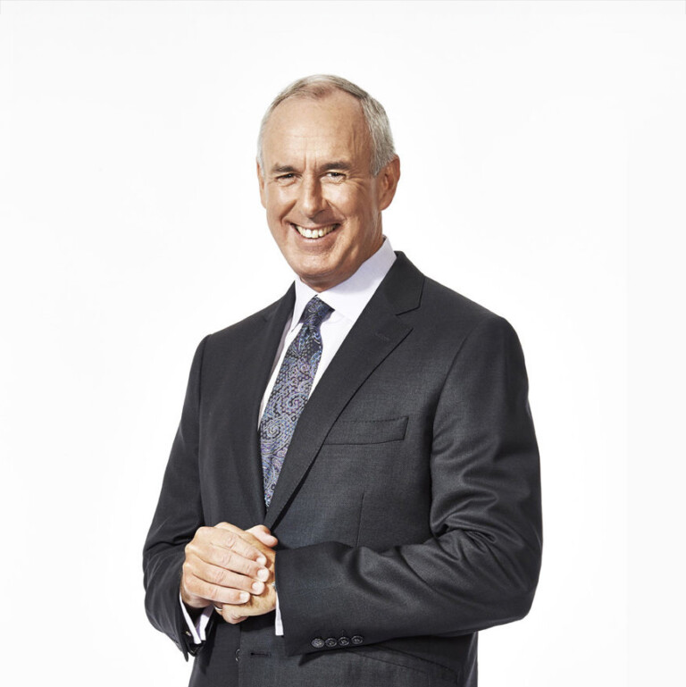Ron MacLean. Headshot of Rob MacLean wearing a grey suit and tie with a white shirt. He is standing with his hands crossed in front of a white background.