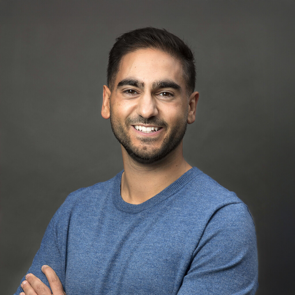 Alex Banayan. Headshot of Alex Banayan wearing a blue top and standing in front of a dark grey background. He is smiling and is crossing his arms.