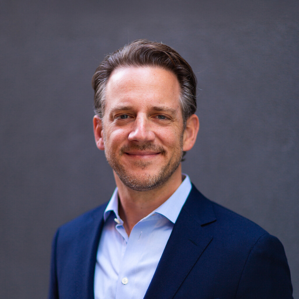 Andrew Kirsch. Headshot of Andrew Kirsch wearing a dark blue suit and a light blue collared shirt. He is posing in front of a dark grey background.