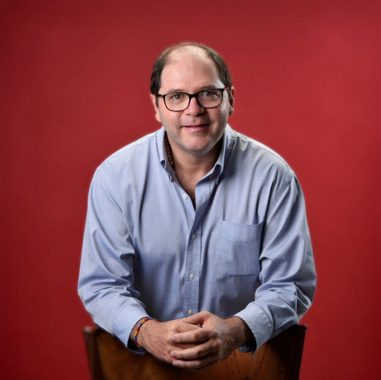 Felipe Gomez. Headshot of Felipe Gomez wearing a light blue collared shirt, leaning over the back of a chair with his hands crossed in front of him. He is standing in front of a red background.