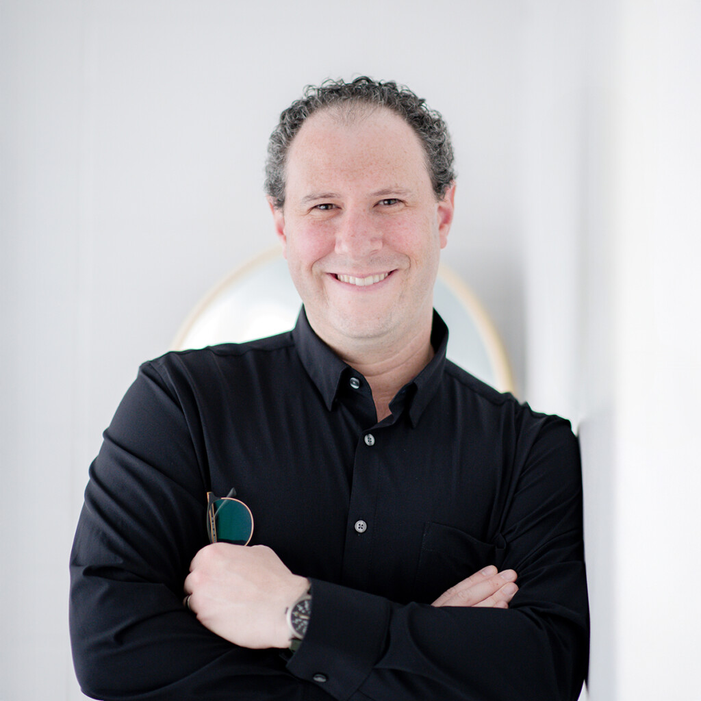 Stephen “Shed” Shedletzky. Headshot of Stephen “Shed” Shedletzky wearing a black button down shirt. He is leaning against a white wall with his arms crossed, holding a pair of sunglasses. He is smiling.