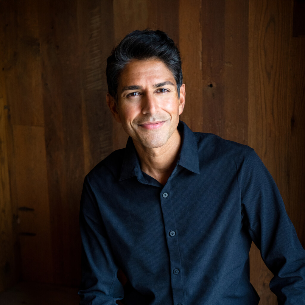 Suneel Gupta. Headshot of Suneel Gupta wearing a dark navy collared shirt and standing in front of a dark wood panel wall.