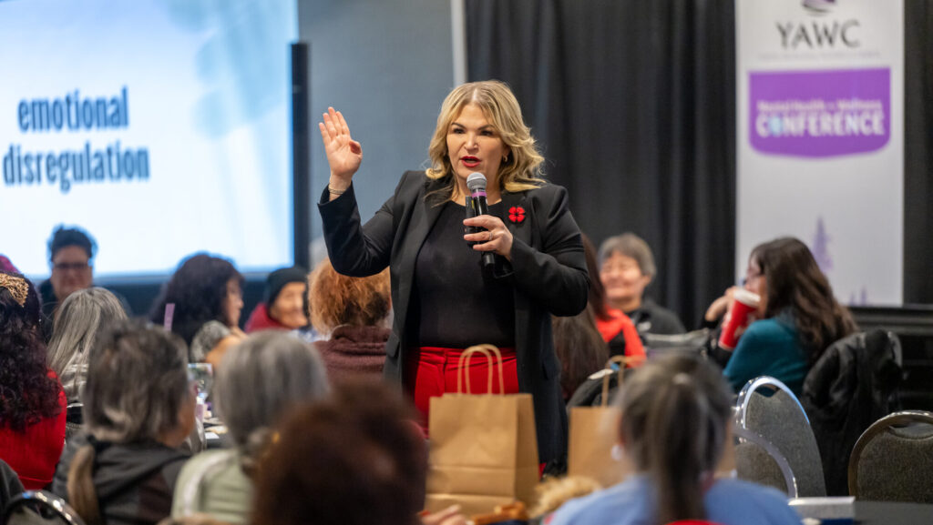Jody Carrington speaking to a audience-filled room. She is holding and speaking into a microphone. She's wearing a black top and blazer and red dress pants.