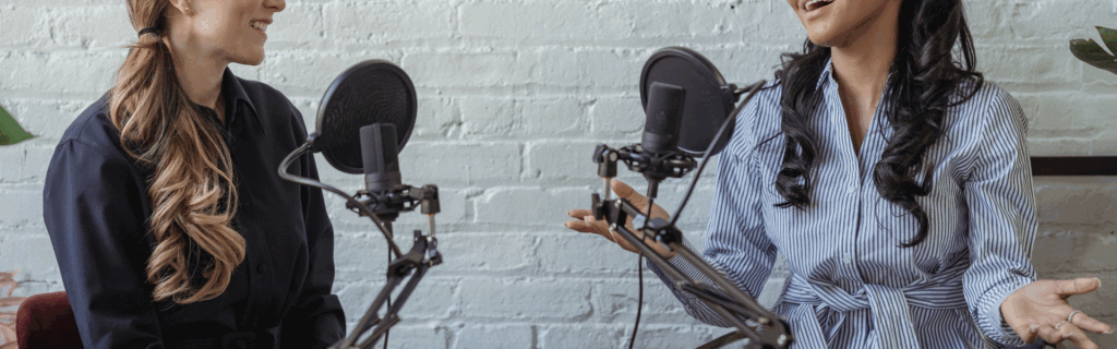 Banner image of two women sitting down at a desk in front of two microphones having a conversation while filming a video or audio podcast.