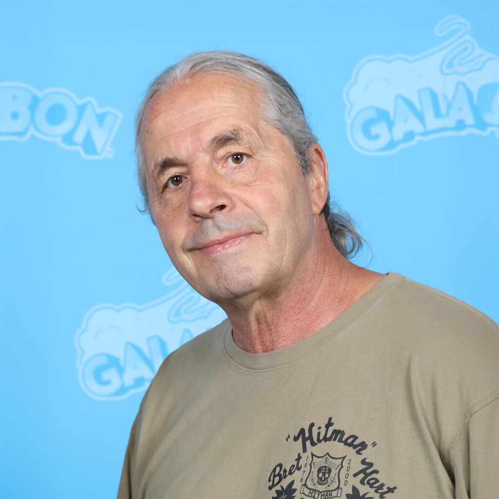 Bret Hart. Headshot of Bret Hart wearing a beige t-shirt and standing in front of a light blue wall.