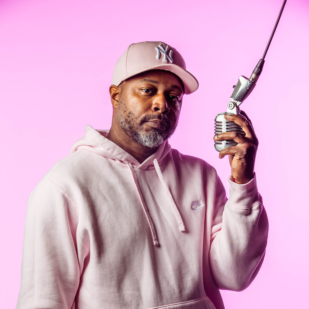 Dwayne Morgan. Headshot of Dwayne Morgan wearing a light pink sweatshirt and cap. He is posing with a microphone and standing in front of a bright pink background.