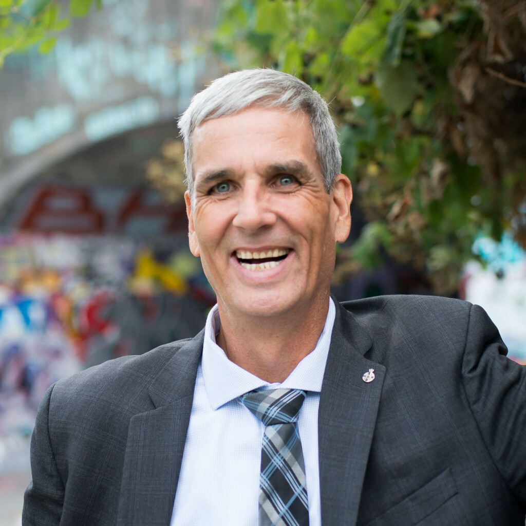 Joe Roberts. Headshot of Joe Roberts wearing a grey suit over a white shirt with a plaid tie. He is smiling and standing outside.