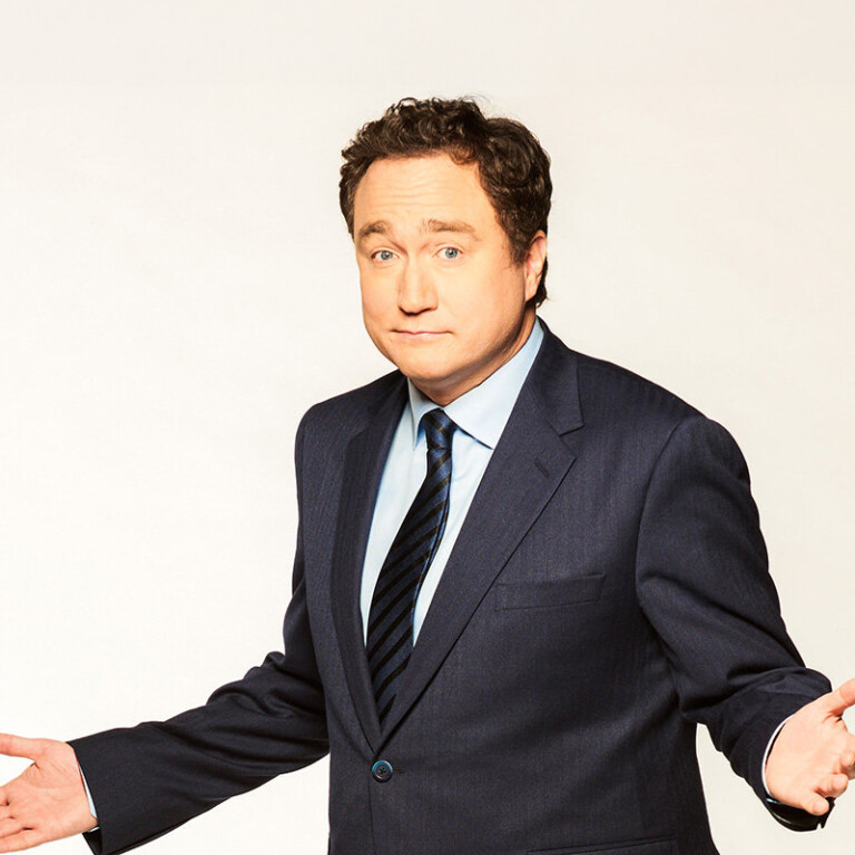 Mark Critch. Headshot of Mark Critch wearing a black suit and tie. He is standing with his arms apart in front of a white background.