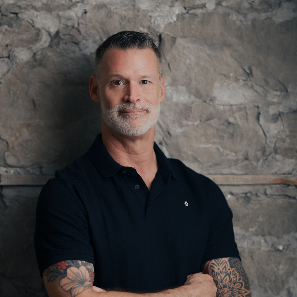 Mark Tewksbury, Headshot of Mark Tewksbury wearing a black short sleeved collared shirt. He is standing with his arms crossed in front of a stone wall.
