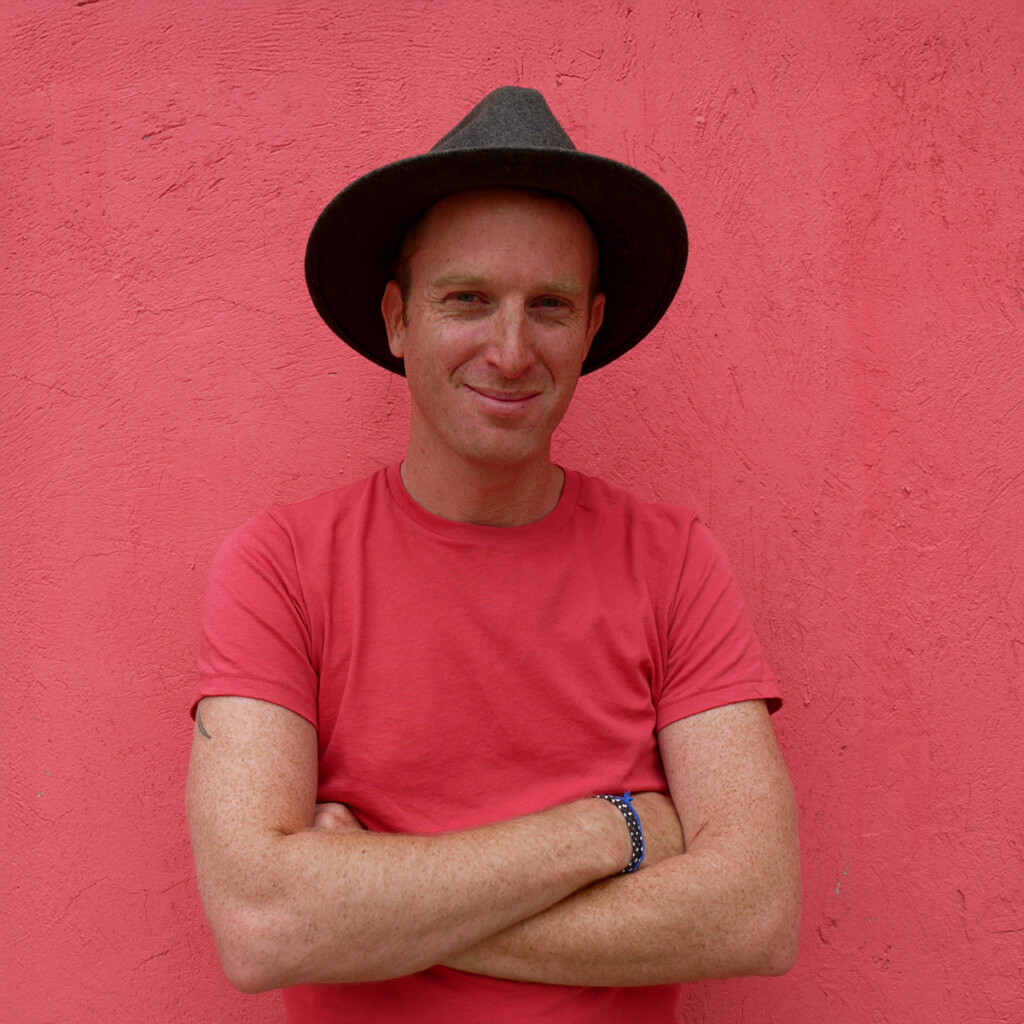 Robin Esrock. Headshot of Robin Esrock. He is wearing a red T-shirt and a black wide-rimmed had. He is crossing his arms and standing in front of a red wall.