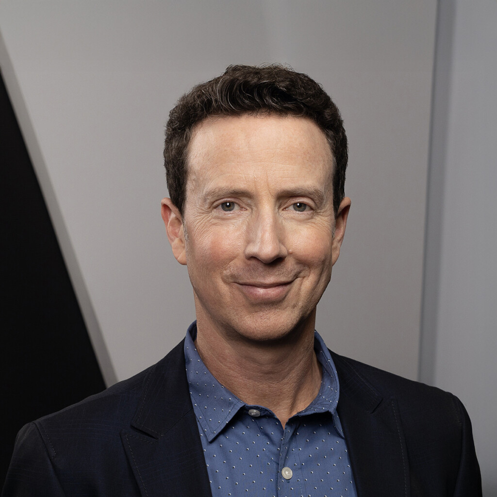 Adam Growe. Headshot of Adam Growe hearing a black suit jacket over a blue collared shirt. He is standing in front of a grey wall.
