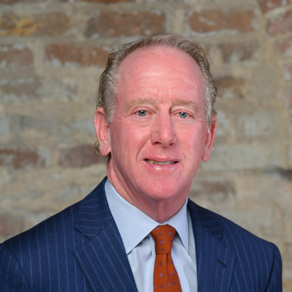 Archie Manning. Headshot of Archie Manning wearing a dark blue suit, light blue shirt and a red tie. He is posing in front of a brick textured background.