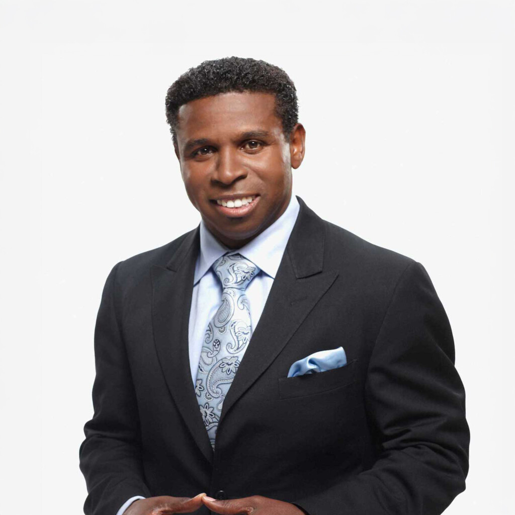 Headshot of Michael Pinball Clemons wearing a black suit with a white shirt and a light blue tie. He is smiling and posing in front of a bright white backdrop.