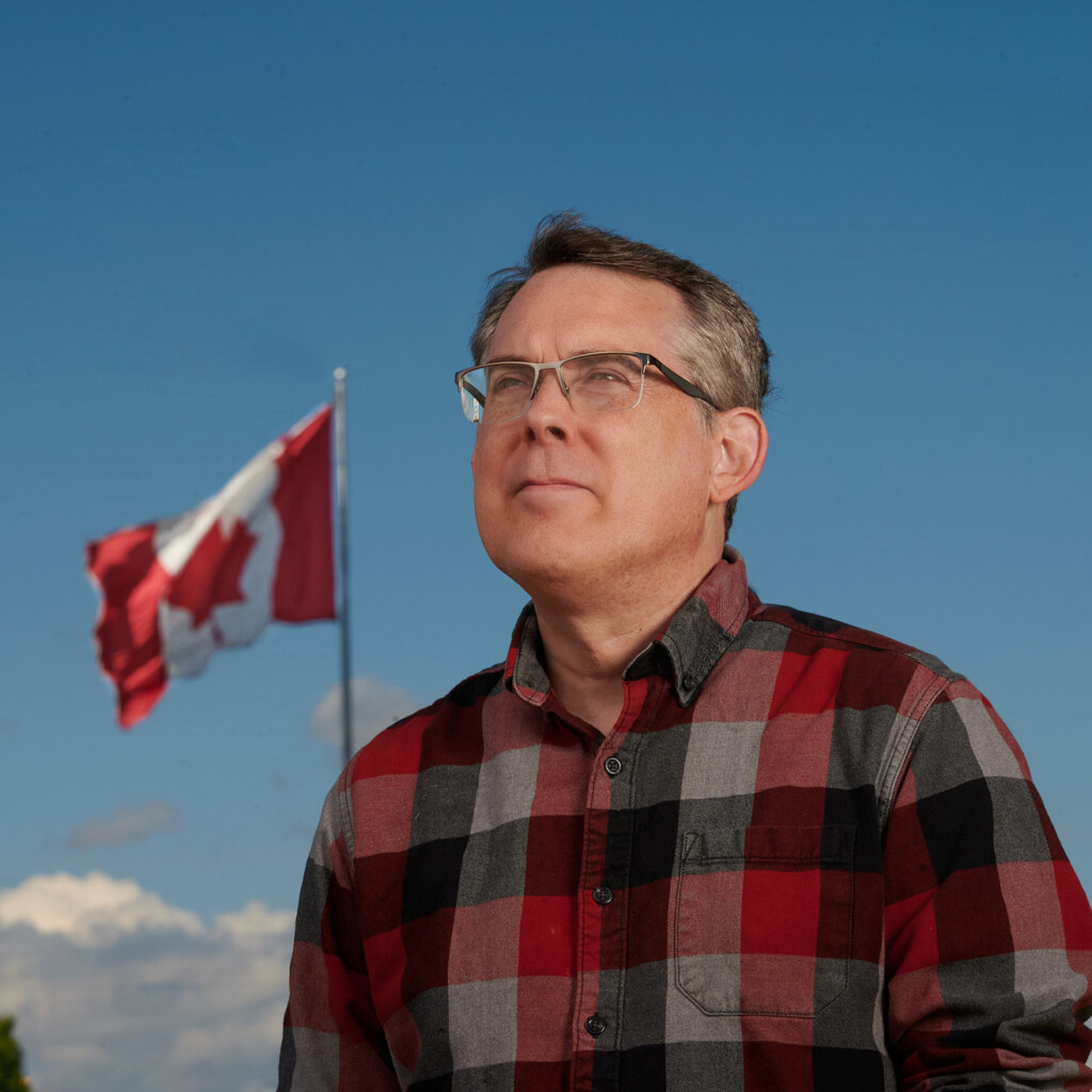 Tod Maffin. Headshot of Tod Maffin wearing a red plaid shirt. He is standing outside, looking sideways with a Canada flag blowing in the wind behind him.