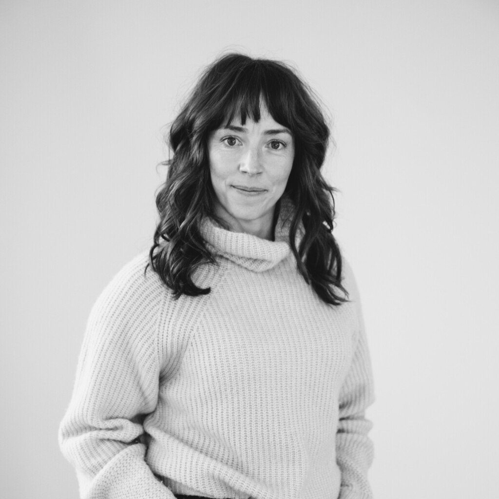 Headshot of Jill Payne. A black and white portrait of Jill standing up with her hands in her front pockets. She has a soft smile and is looking at the camera.