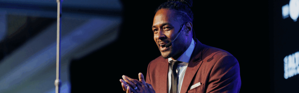 Jesse Lipscombe onstage hosting an event. He is wearing a maroon suit.