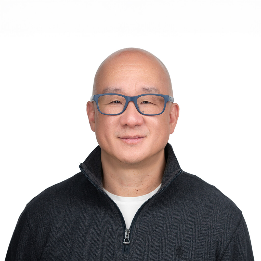 Rocky Ozaki. Headshot of Rocky Ozaki wearing a black zip-up sweater over a white t-shirt. He is posing in front of a bright white backdrop.