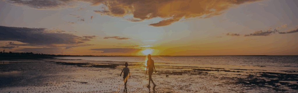 Two people walking on a beach next to the shoreline and as the sun sets in the background creating an orange-coloured sky.