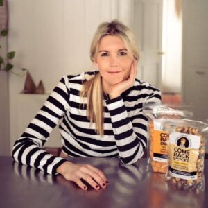 Photo of keynote speaker, Emily O'Brien, smiling, and sitting at a kitchen table. She is wearing a striped, black and white long-sleeve shirt. Two bags of her Comeback Snacks are on the table in front of her.
