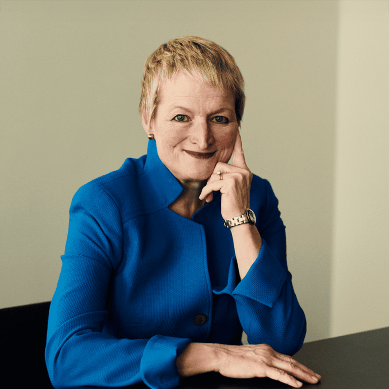 Rita McGrath. Headshot of Rita McGrath wearing a blue collared jacket. She's sitting down and posing with one hand on her cheek. She's in front of a grey/green wall.
