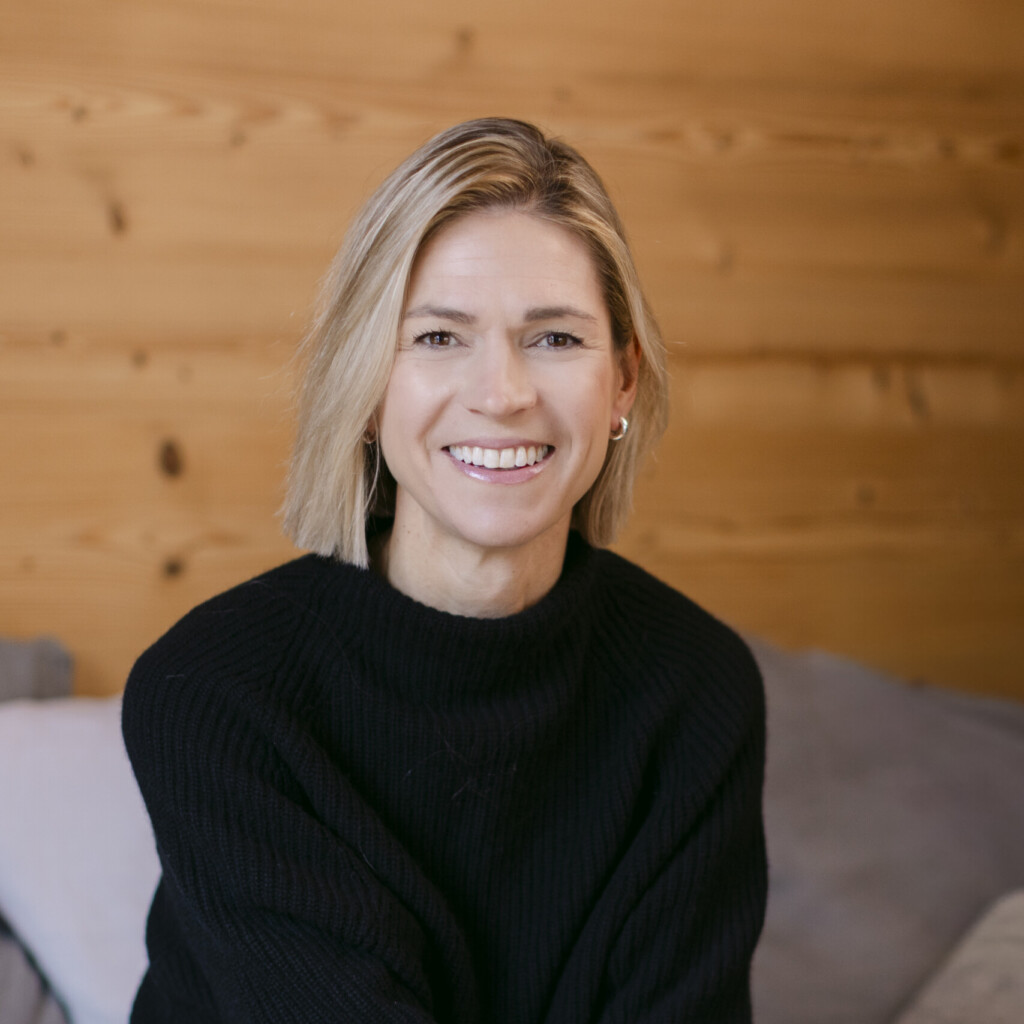 Headshot of speaker, Stephanie Case, wearing a black long-sleeve shirt and white pants. She is sitting on a grey couch, and smiling at the camera.