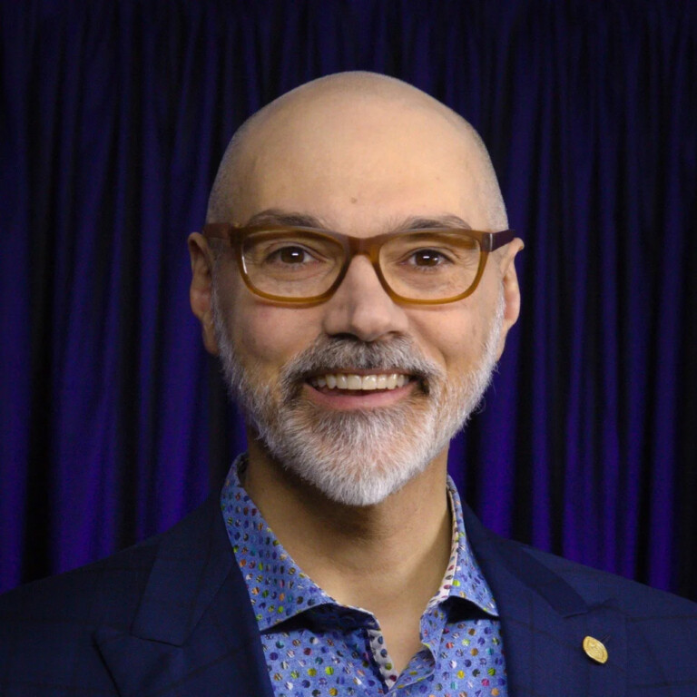 Dan Trommater. Headshot of Dan Trommater wearing a blue jacket over a blue collared shirt. He is posing in front of a dark blue curtain.
