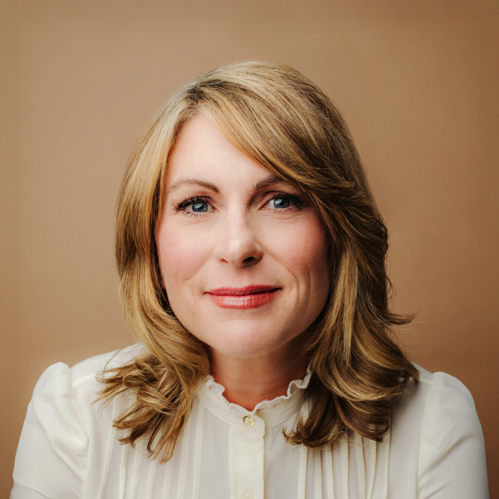 Jennifer Moss. Headshot of Jennifer Moss wearing a white blouse and posing in front of a brown gradient background.