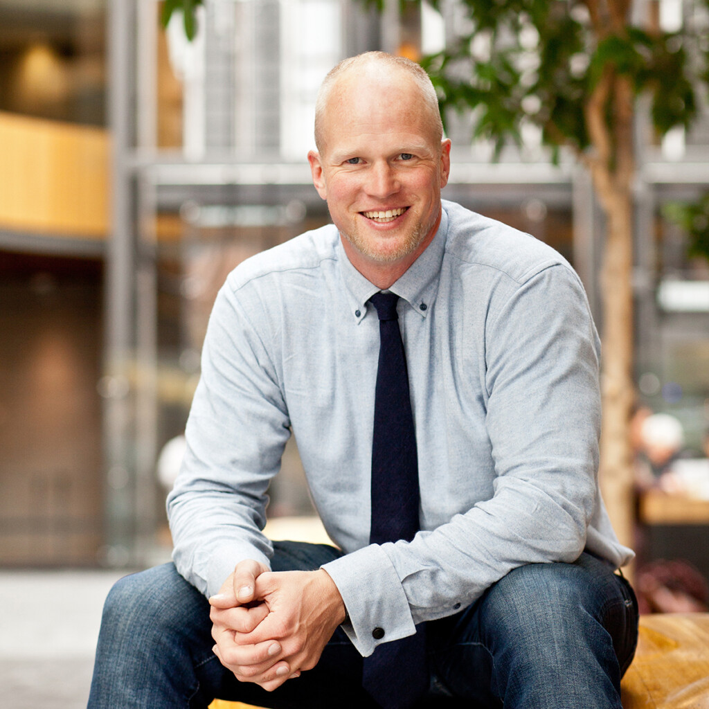 Adam Kreek. Headshot of Adam Kreek wearing a light blue shirt and black tie. He is sitting down outside.