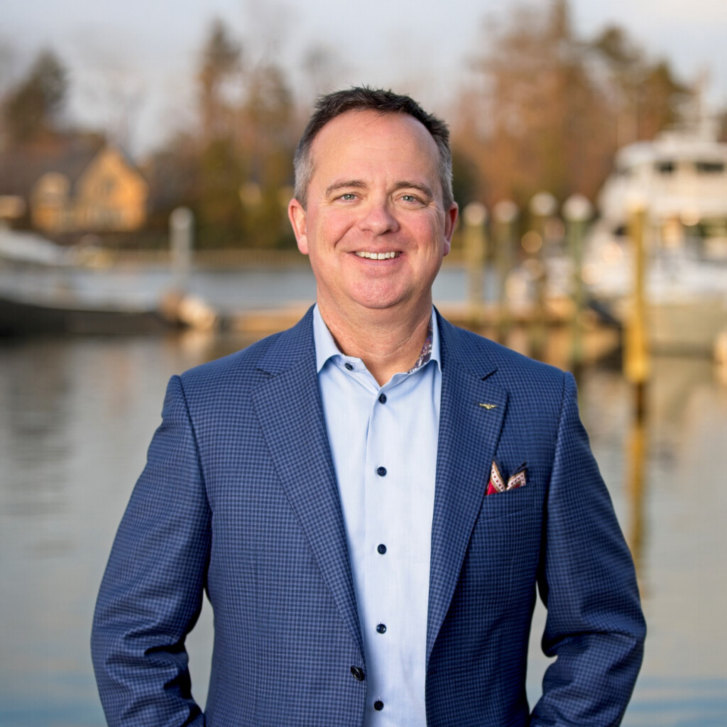 Jack Becker. Headshot of Jack Becker wearing a blue suit over a light blue collared shirt. He's posing outside on a boat dock.
