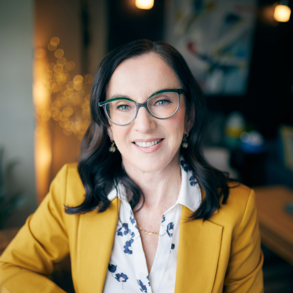 Dr. Jane Barratt. Headshot of Jane Barrat wearing a yellow blazer over a white blouse. She's wearing glasses and sitting inside with a blurred background.
